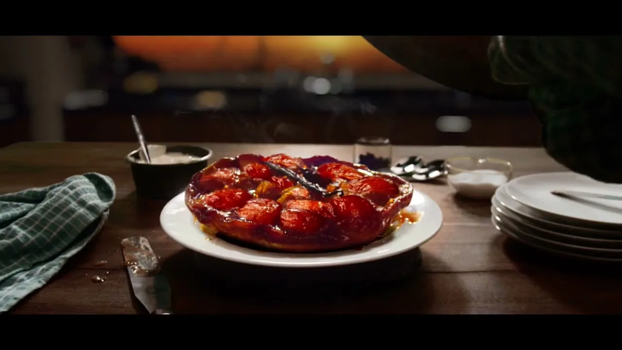 A freshly baked tarte tatin with steam rising is placed on a wooden table, with a person's arm partially visible on the right.