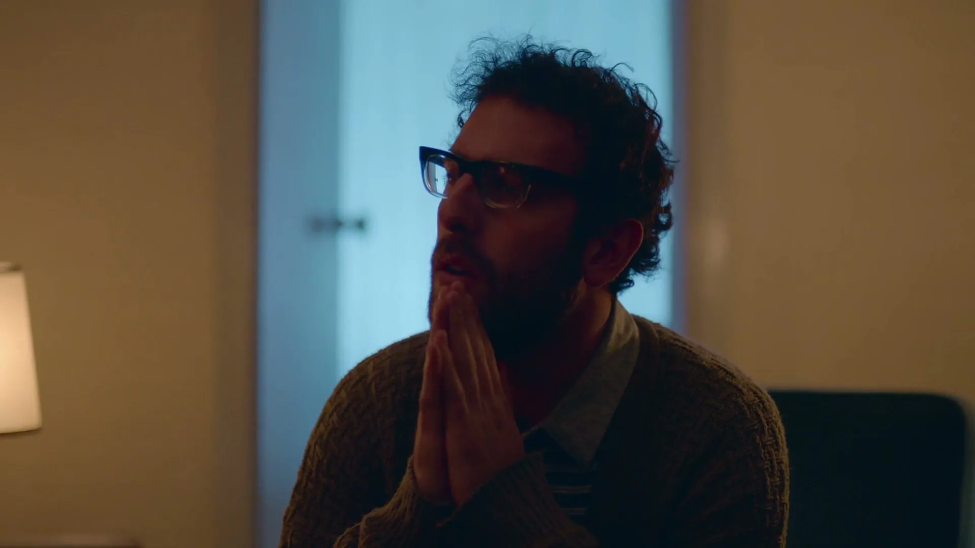 A man with curly hair and glasses sits with his hands clasped, looking up and to the side in a dimly lit room.