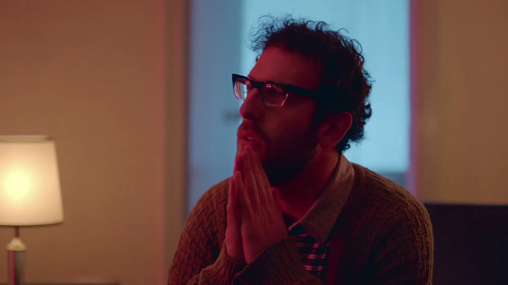 A man with curly hair and glasses sits with his hands clasped, looking up and to the side in a dimly lit room.