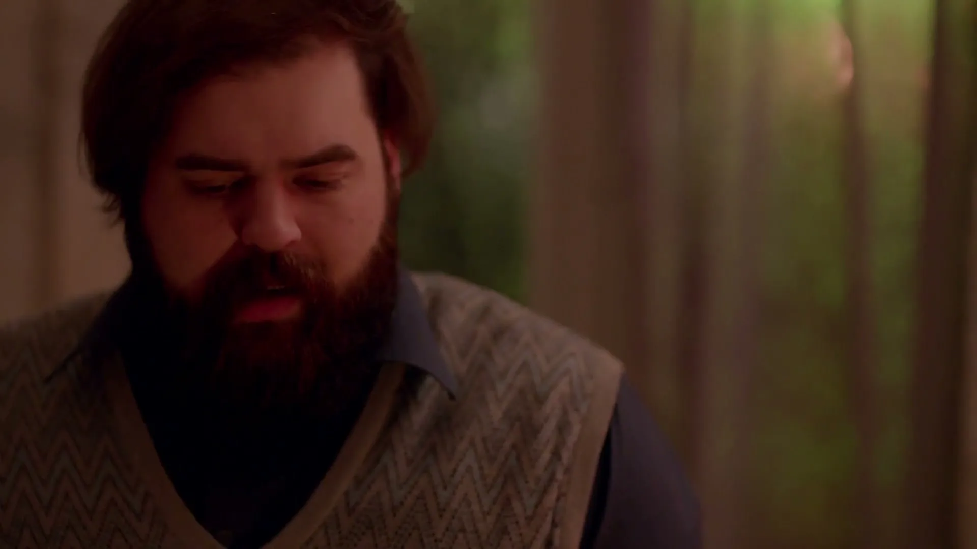 A man with a beard and a sweater vest looks down and then closes his eyes in frustration in an interior setting.