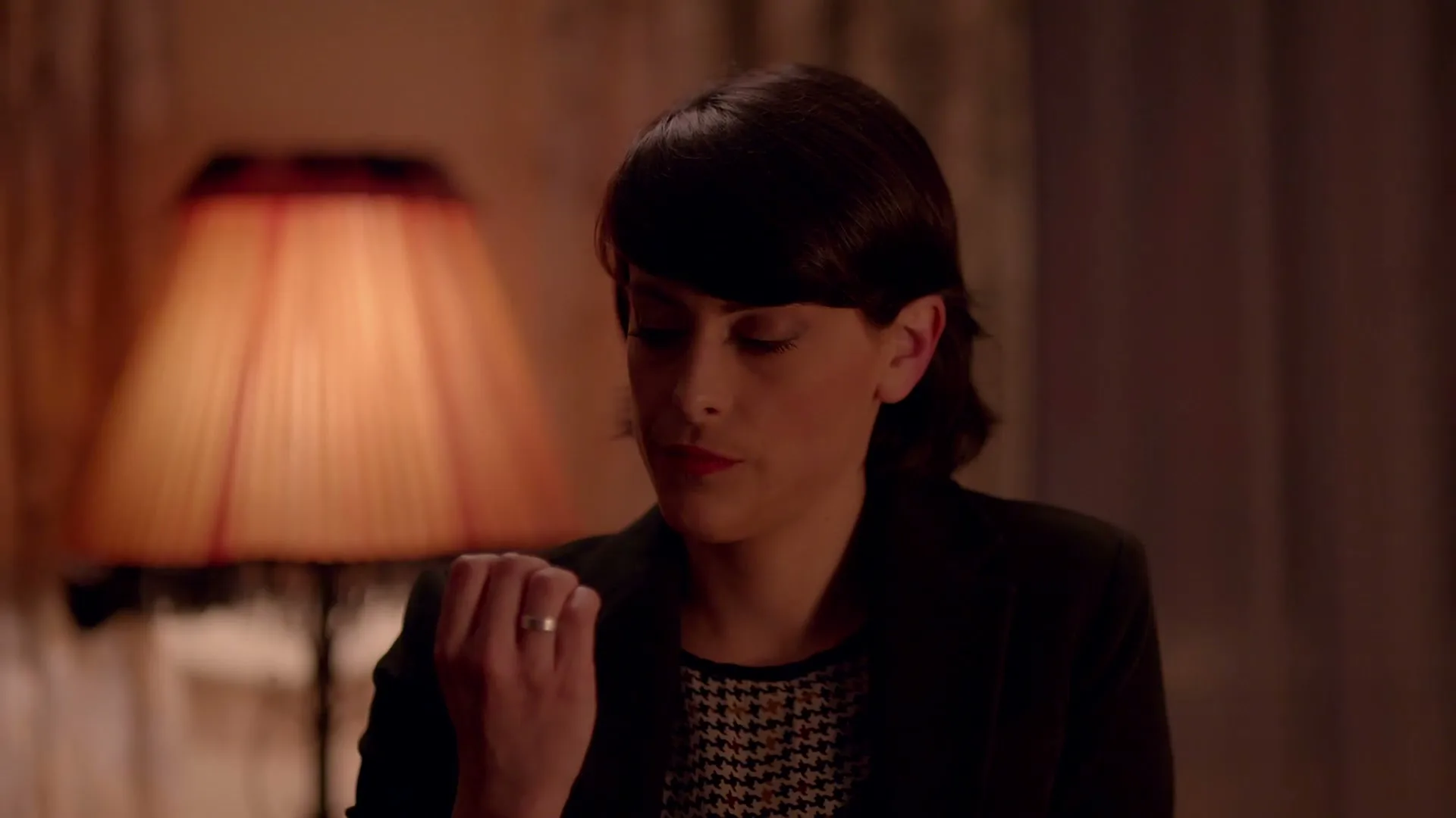A woman with short dark hair, wearing a black blazer and houndstooth top, looks down at her hand in a dimly lit room.