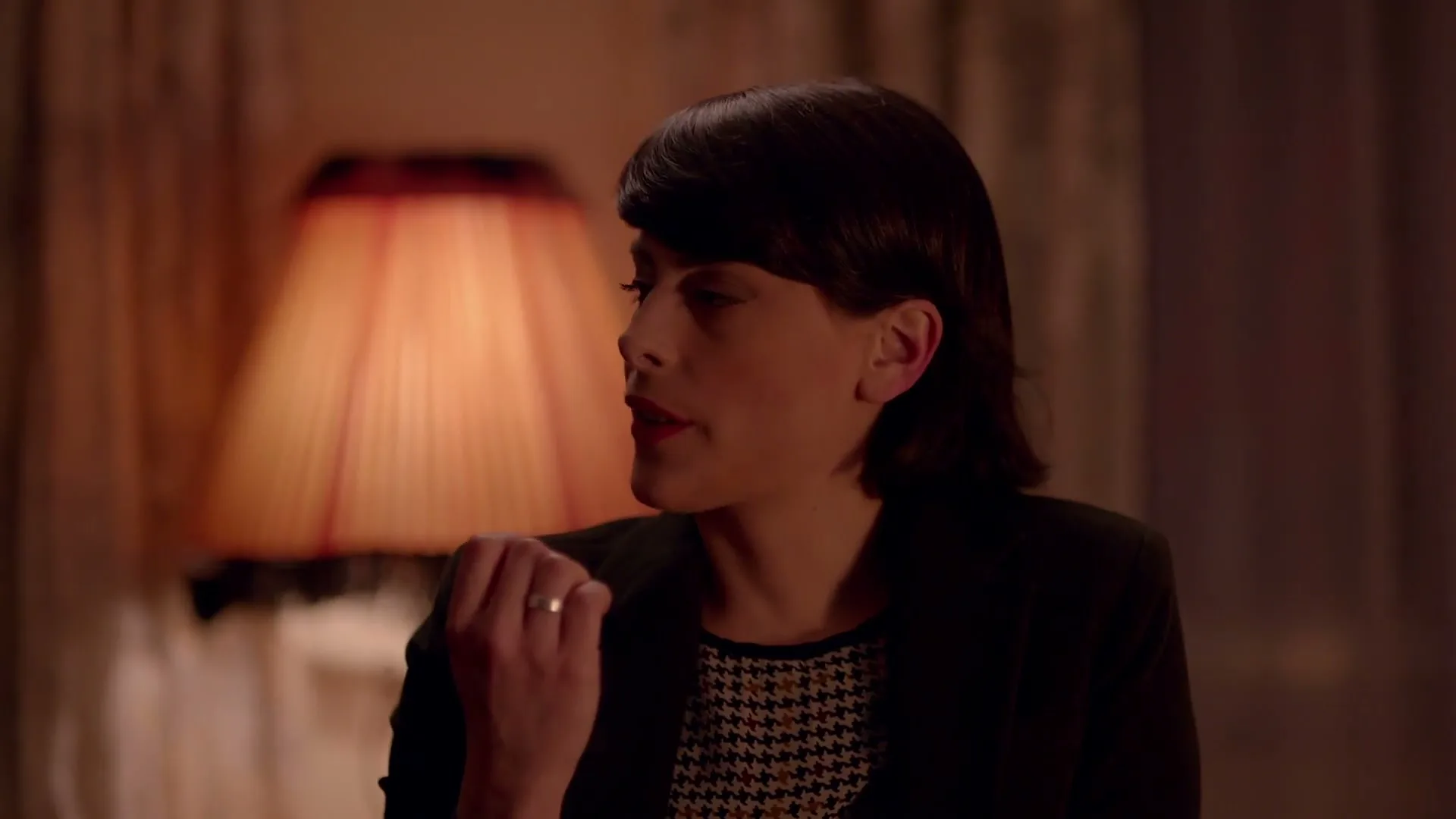 A woman with short dark hair and a houndstooth top talks to someone off-screen in a dimly lit room with a lamp.