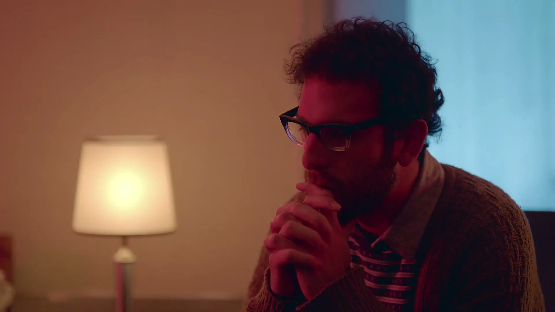 A man with curly hair and glasses sits with his hands clasped, looking pensive in a room with warm and cool lighting.