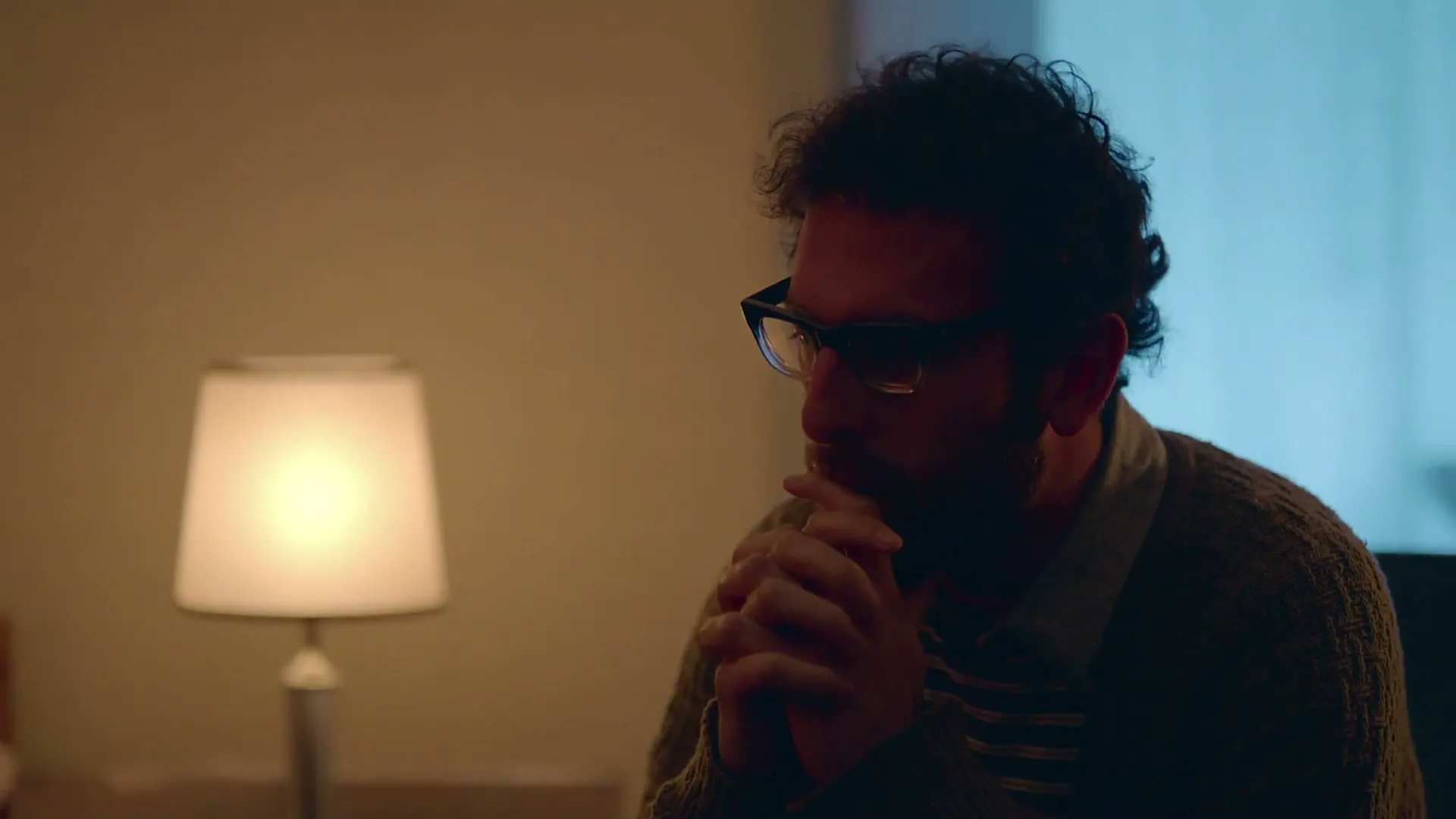 A man with curly hair and glasses sits with his hands clasped, looking pensive in a dimly lit room with a lamp beside him.