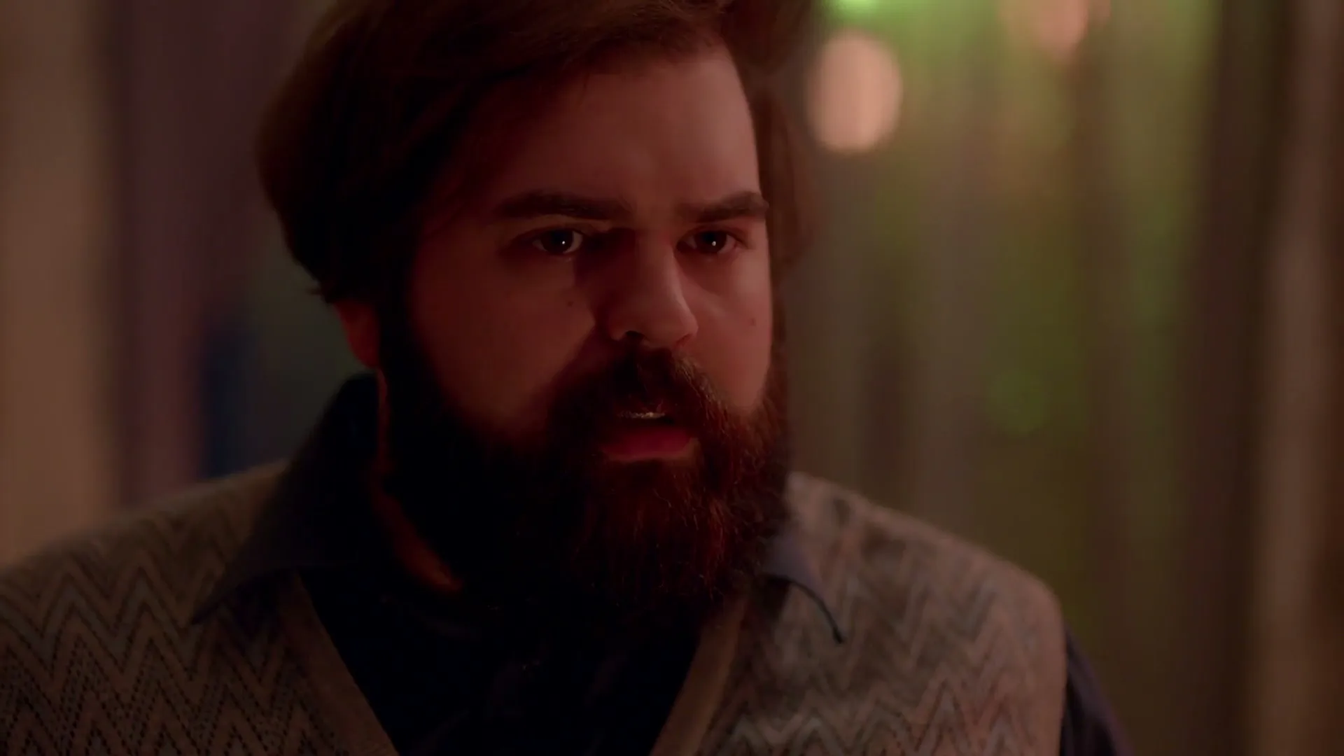 A man with a beard and a patterned vest looks down and then up with a concerned expression in an interior setting.