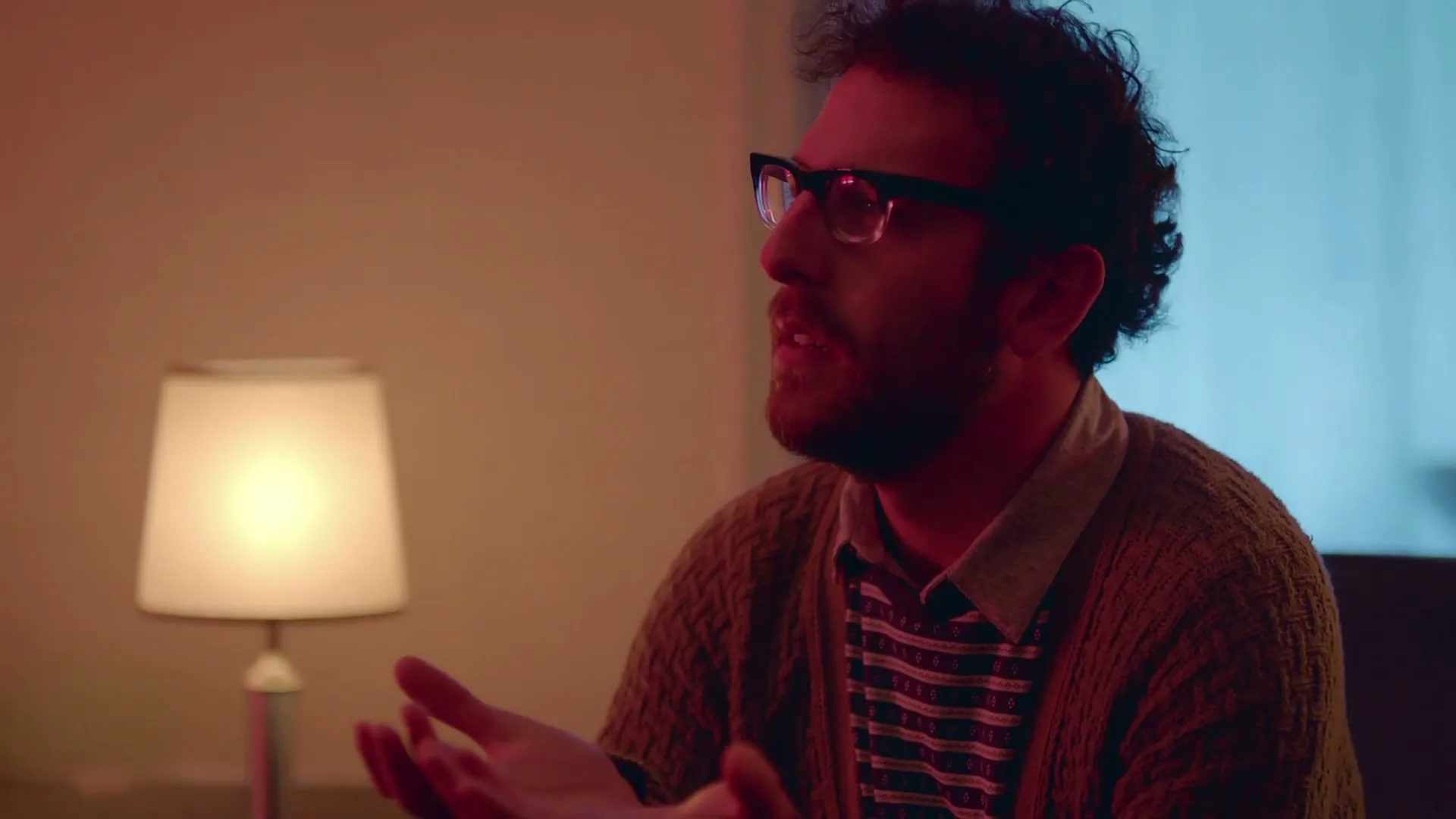 A man with curly hair and glasses, wearing a cardigan, gestures with his hands while talking in a dimly lit room.