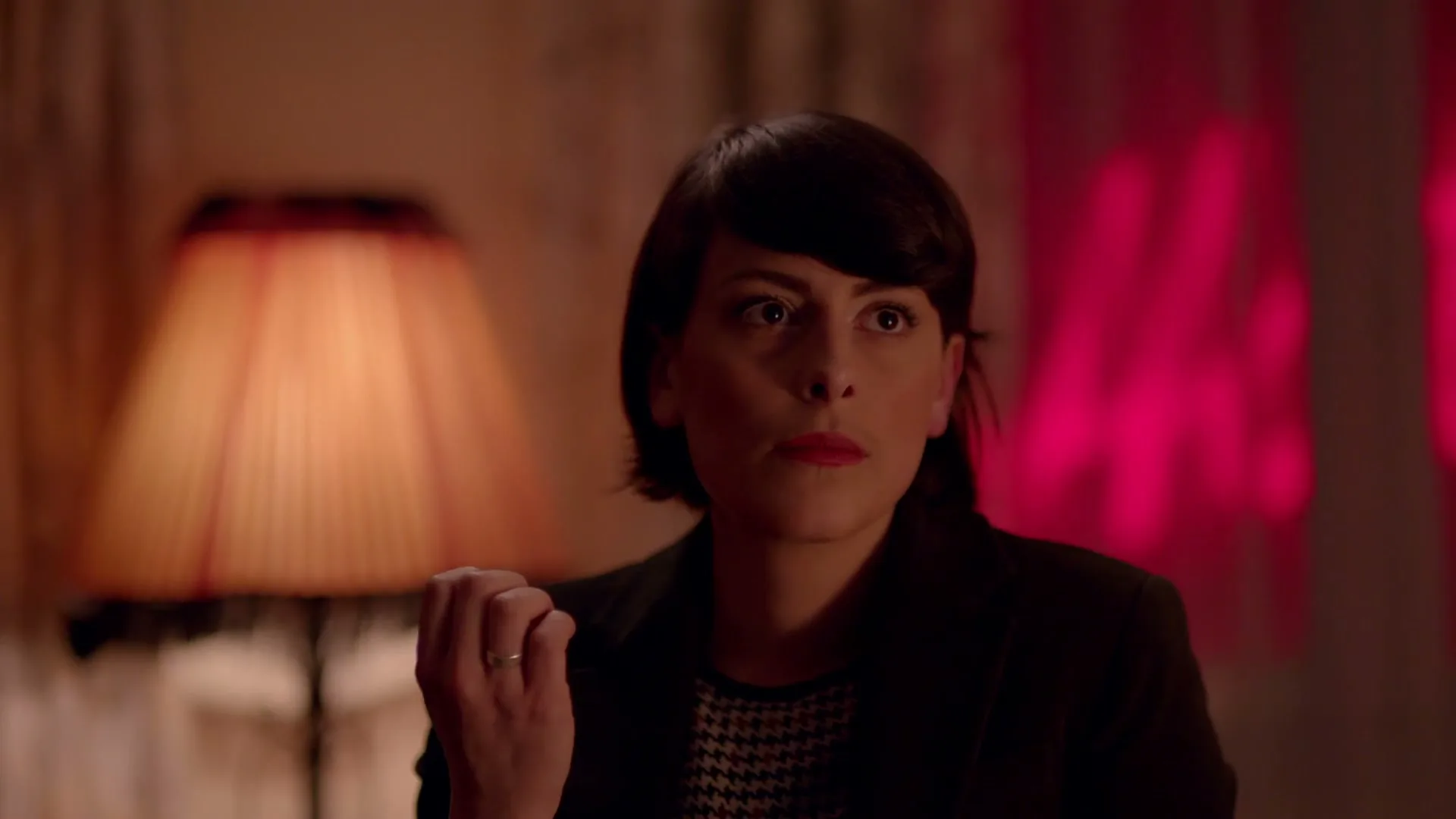 A woman with short dark hair and red lipstick looks intently to the right, with a lamp and red light in the background.
