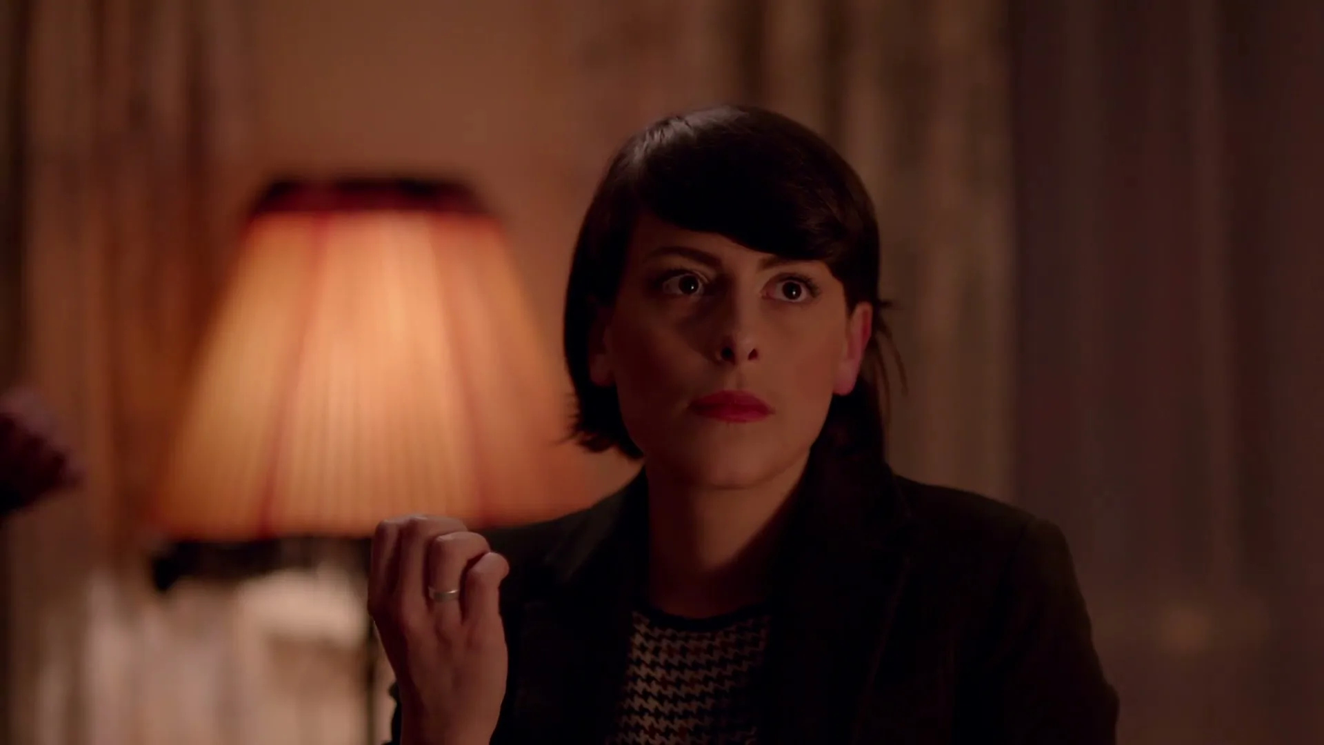 A woman with short dark hair and red lipstick looks intently at something off-screen in a dimly lit room with a lamp.