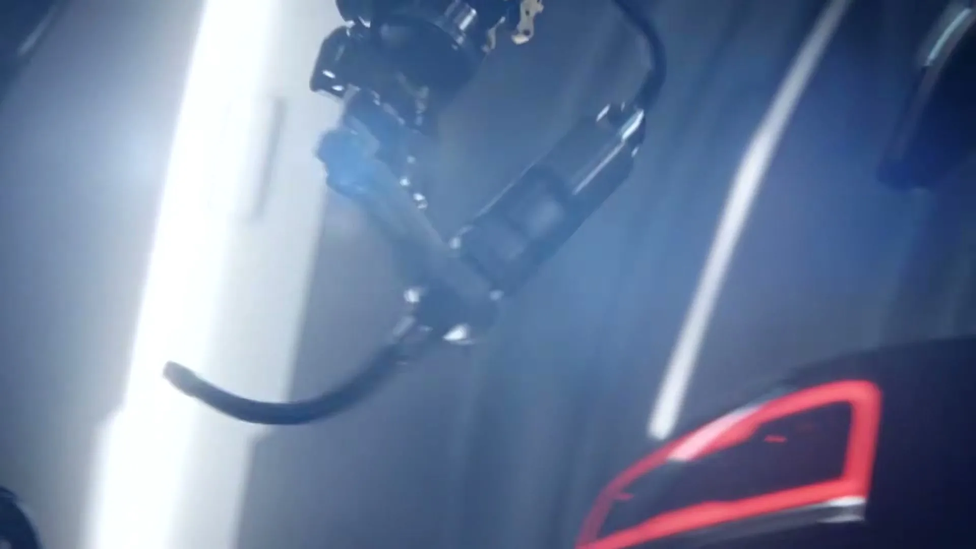 A robotic arm with a drill attachment moves towards the rear of a dark car with illuminated taillights in a brightly lit, futuristic factory.