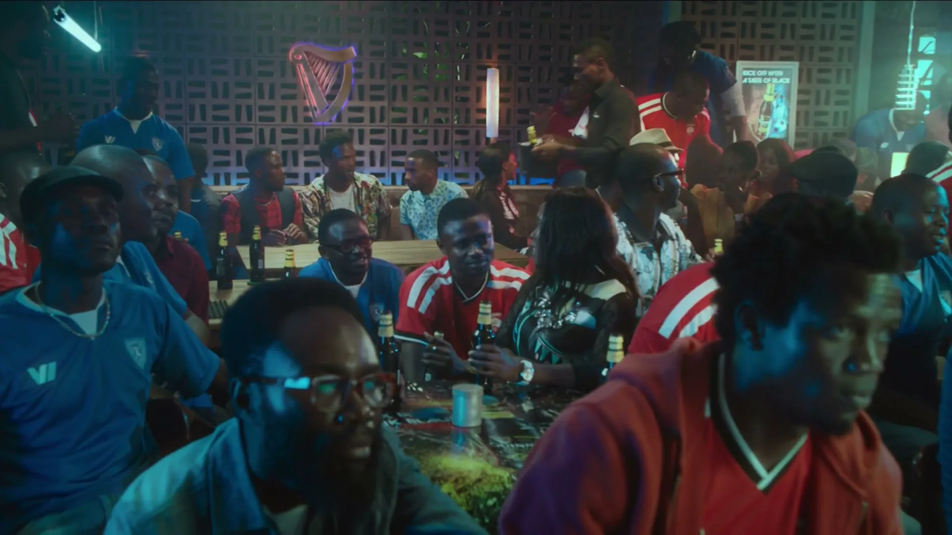 A diverse group of people, mostly men, are gathered in a dimly lit bar, watching something intently.