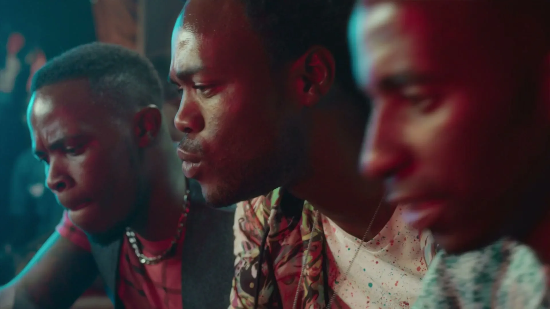 Three young men with dark skin are sitting in a dimly lit, crowded club, looking intently at something off-screen.