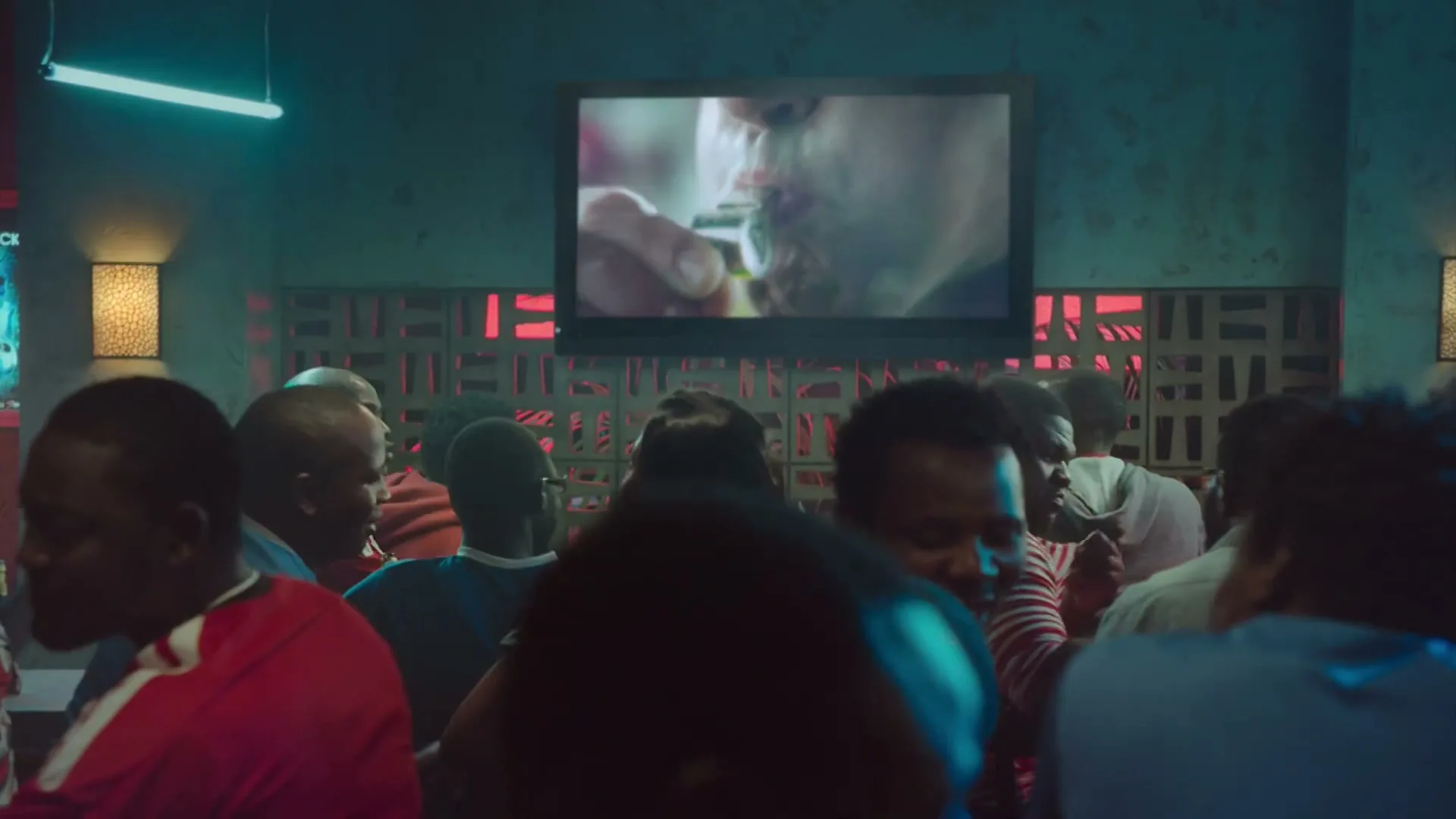 A diverse group of people watch a soccer referee blow a whistle on a TV screen in a dimly lit bar.