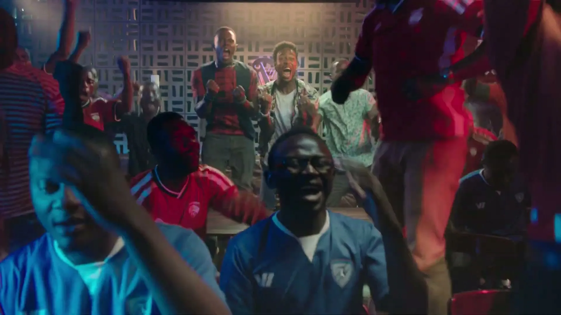 A group of young adults, mostly male, cheer excitedly in a dimly lit bar, celebrating a sports event.