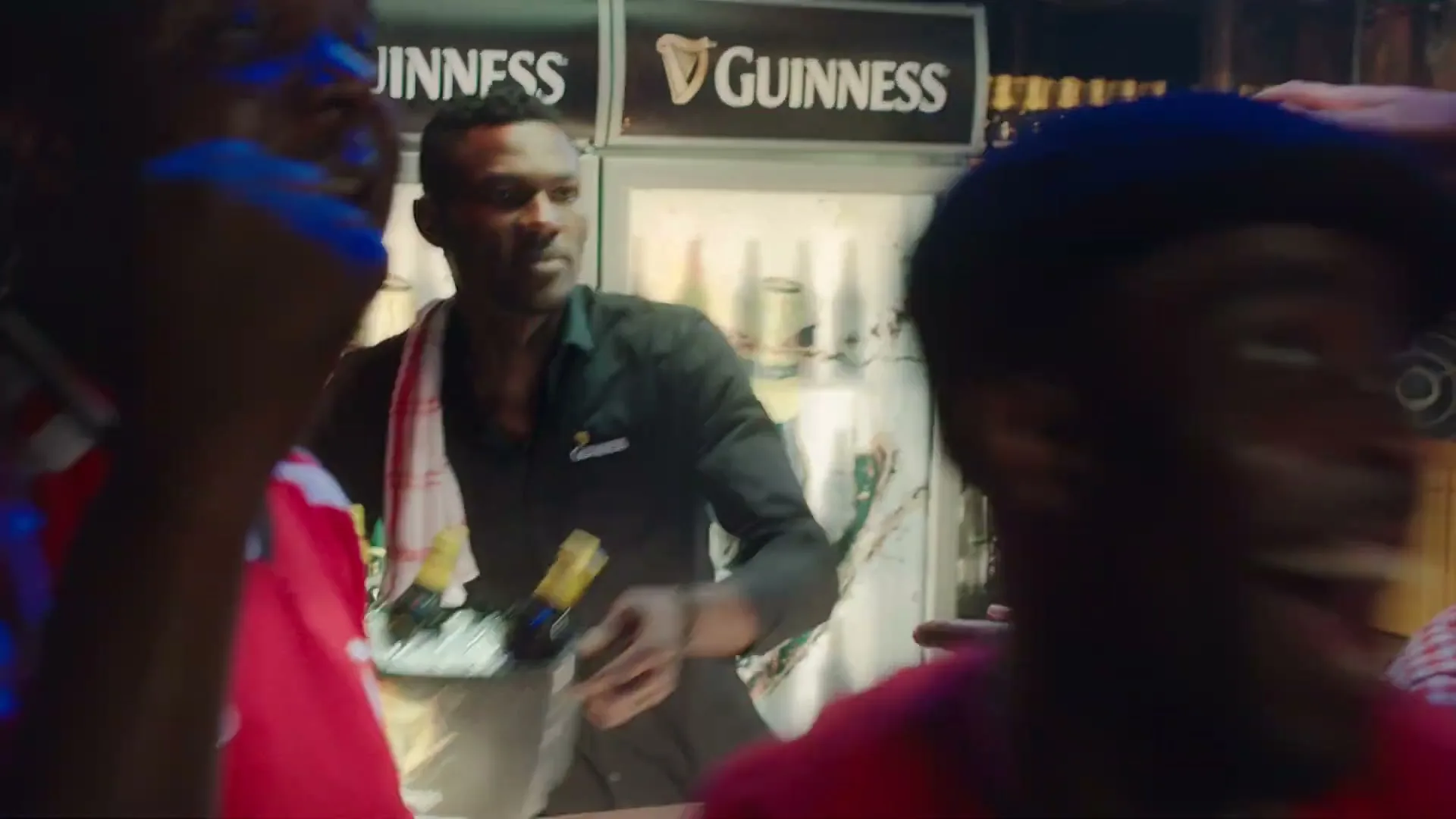 A bartender in a black shirt and white towel serves a bucket of Guinness beer bottles to an excited customer in a lively bar.