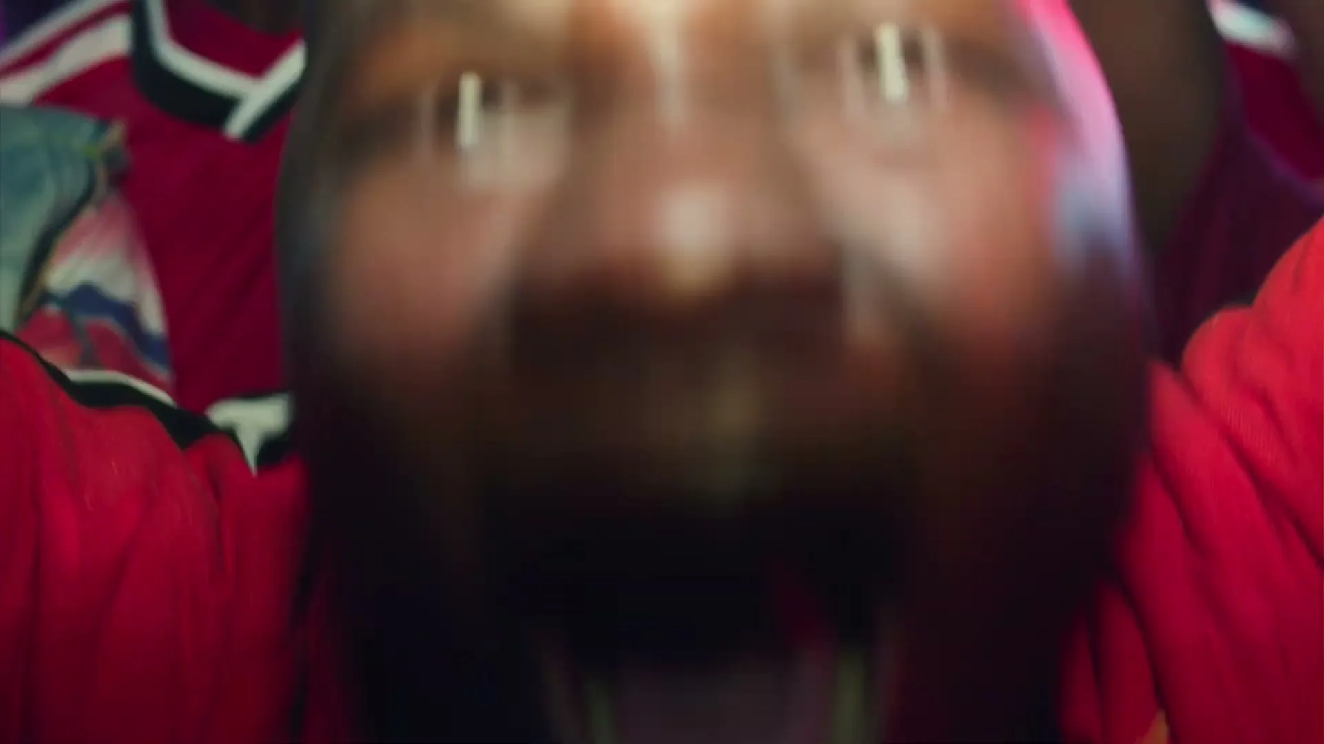 A man in a red shirt screams with his mouth wide open, seen from a low angle in a dark, vibrant setting.