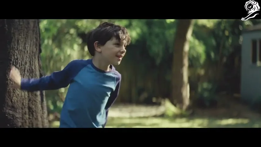 A young boy in a blue shirt counts to himself while playing hide-and-seek in a sunny backyard.