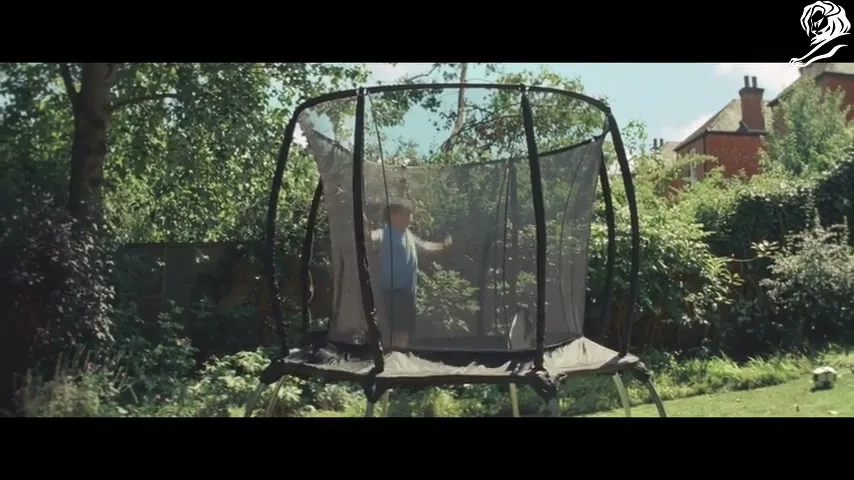 A young boy in a blue t-shirt jumps on a trampoline in a sunny suburban backyard, surrounded by green trees and bushes.