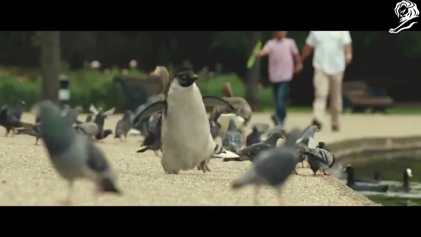 A CGI penguin stands among pigeons and geese by a pond in a park, while a boy and man walk hand-in-hand in the background.