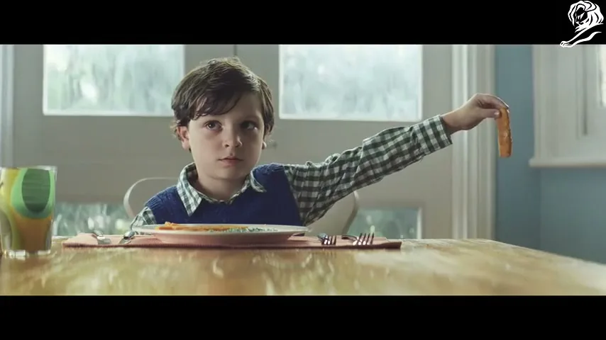 A young boy with dark hair and a blue vest sits at a dining table, holding up a carrot stick for a penguin toy to eat.