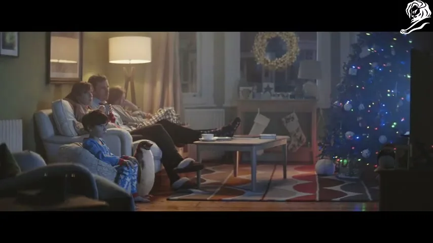 A family of four, including a young boy in pajamas with a toy penguin, watches TV together in a cozy living room decorated for Christmas.