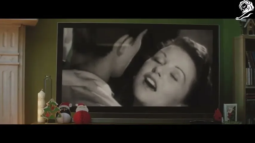 A black and white movie scene of a man and woman kissing is shown on a television screen, surrounded by Christmas decorations in a living room.