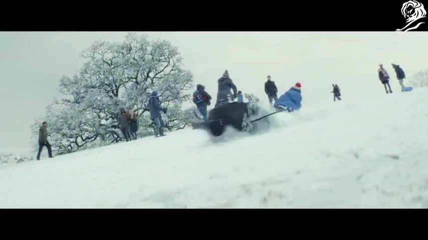A young boy in a blue coat and red hat sleds down a snowy hill with a toy penguin, while other people watch and play in the background.