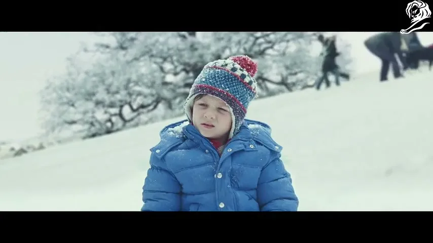 A young boy in a blue winter coat and colorful hat stands in a snowy field, looking thoughtful as people and a dog play in the background.