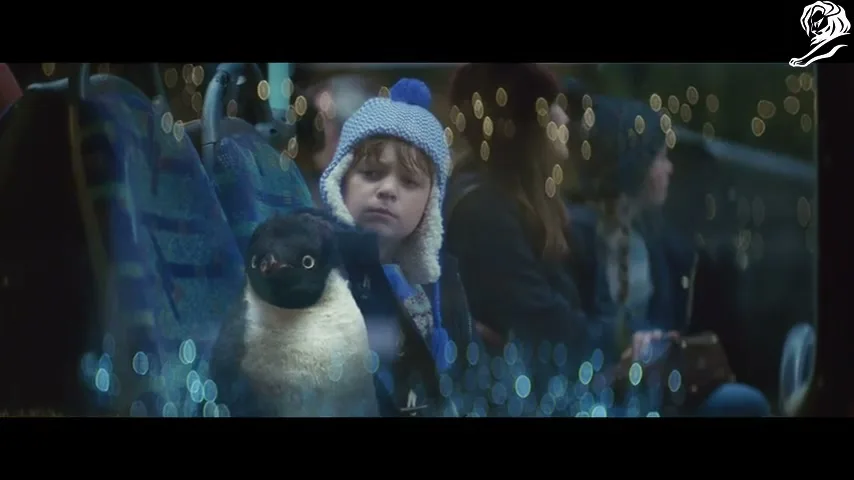 A young boy in a blue hat sits on a bus with a toy penguin, looking out the window at blurry lights.