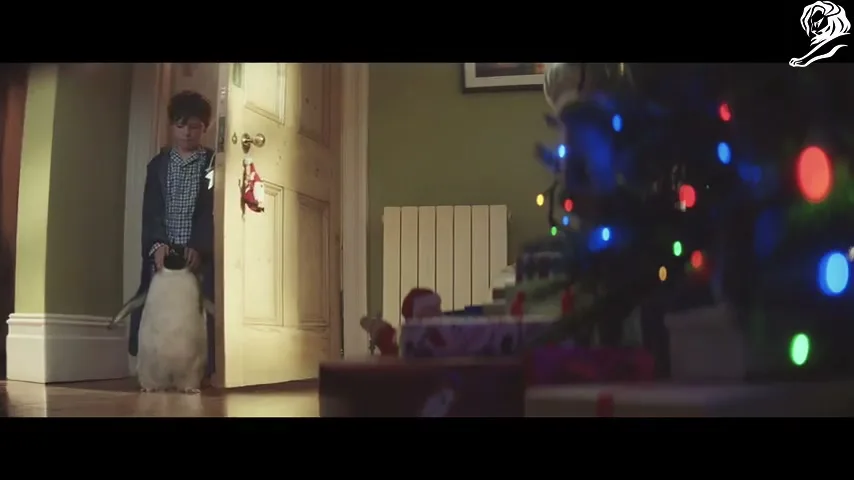 A young boy in pajamas holds a penguin toy, walking through a doorway into a living room with a decorated Christmas tree.