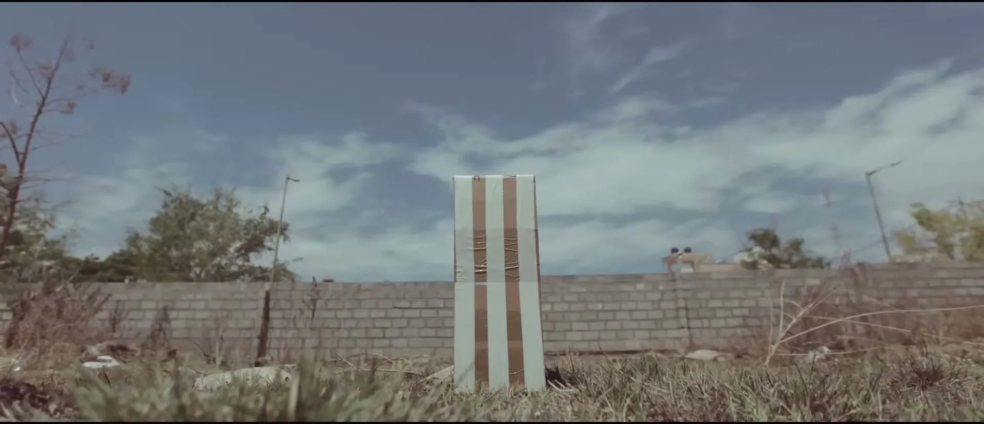 A makeshift cricket wicket, made from cardboard and tape, stands in a grassy, overgrown field under a bright, cloudy sky.
