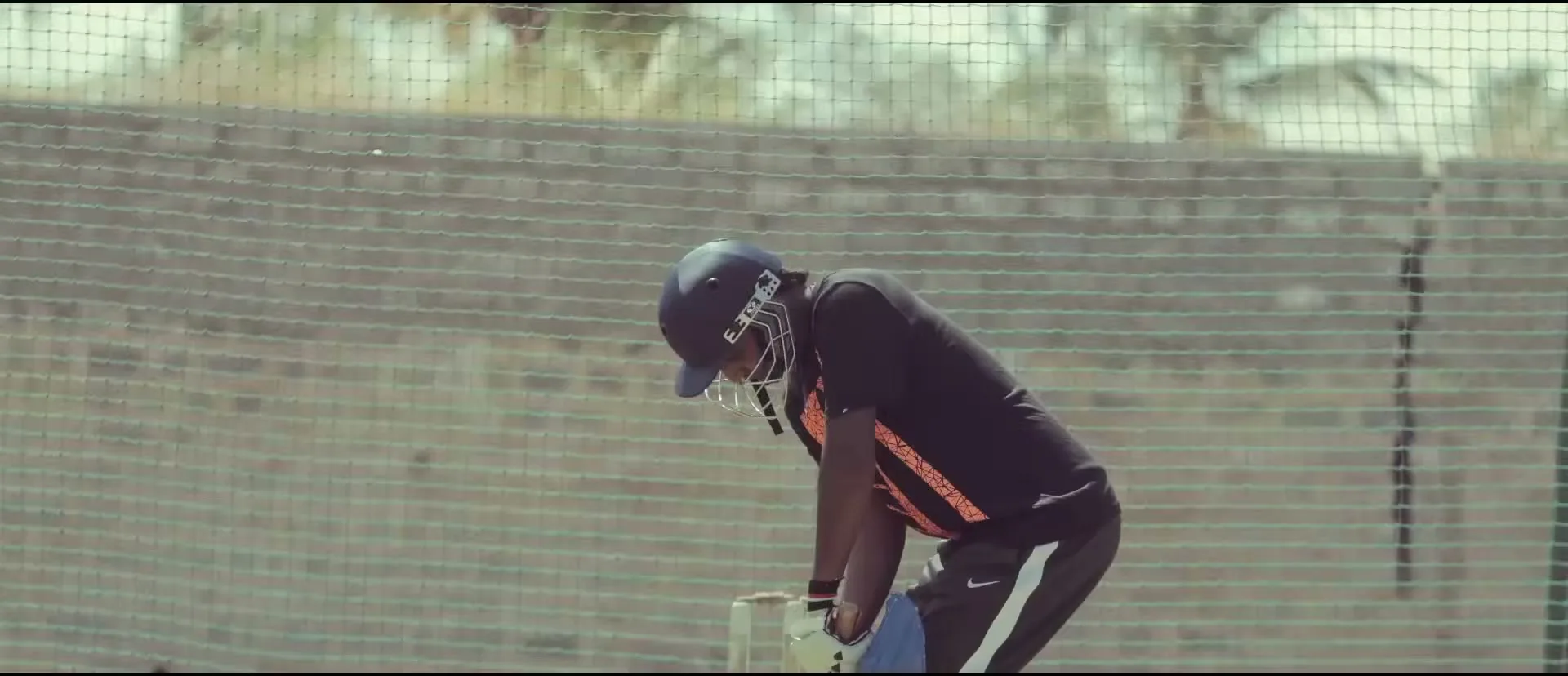 Two young boys play cricket on a dusty field, one batting and the other fielding, with buildings in the background.