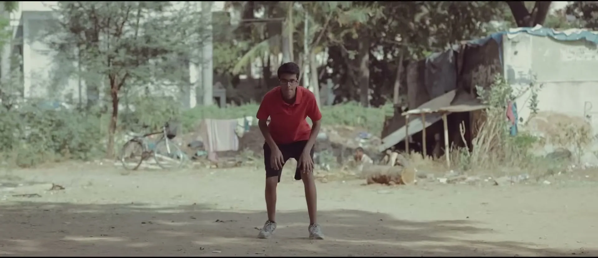 A young boy in a red shirt and black shorts practices cricket in a dusty, outdoor area with makeshift shelters in the background.