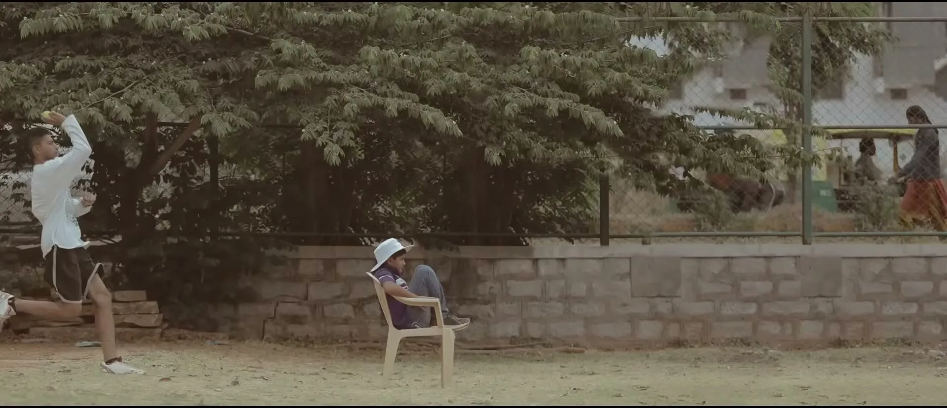 A young male cricketer in white and black uniform bowls a ball on a dirt field, while a child in a white hat sits on a chair watching.