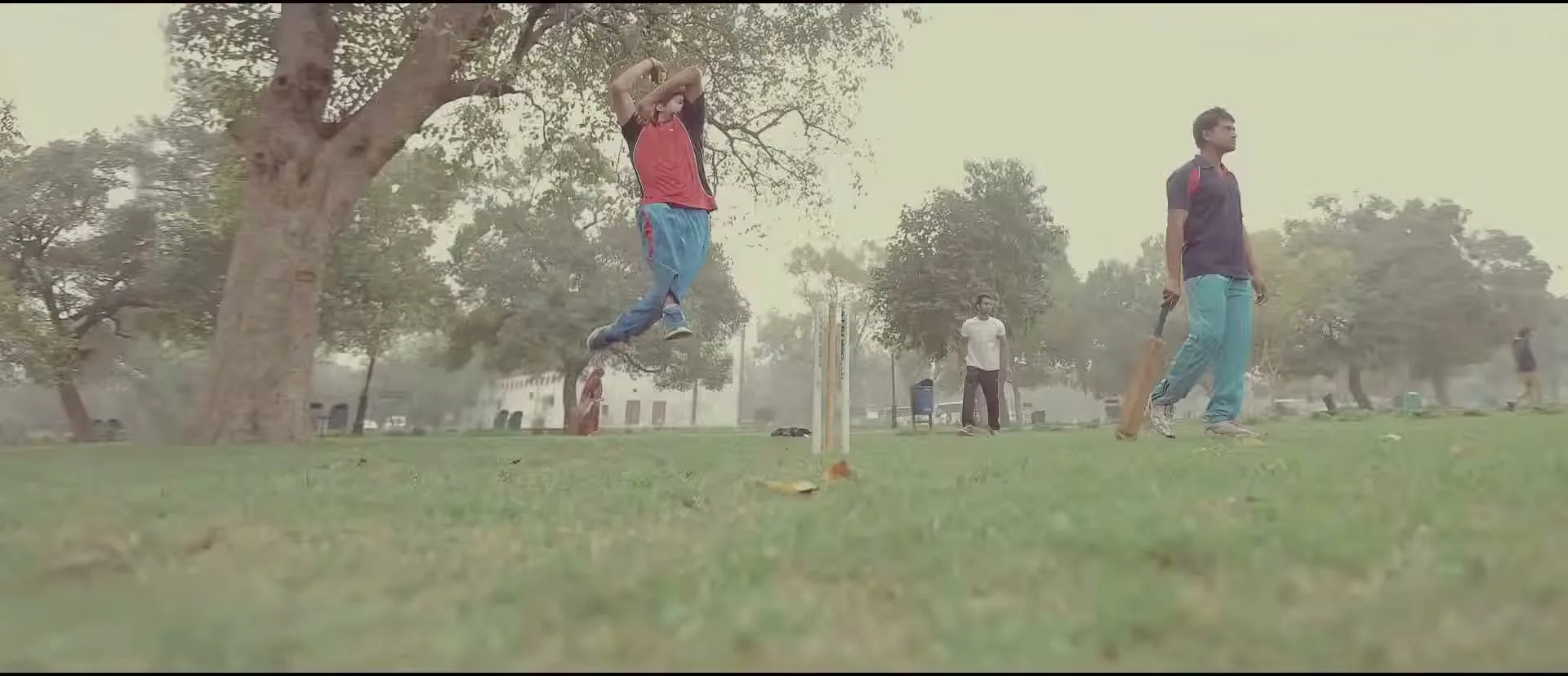 A male cricketer in a blue shirt and shorts jumps high in the air, holding a cricket bat, in a narrow, trash-strewn alleyway.