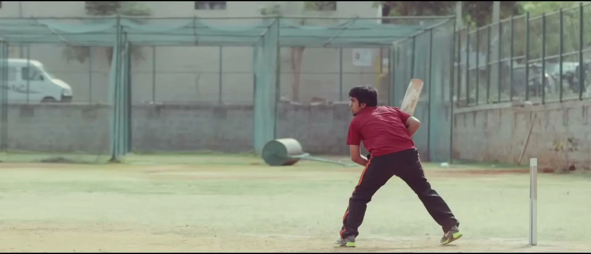 A young male cricketer in a red shirt and black pants swings a bat on a dusty field with a green net in the background during the day.
