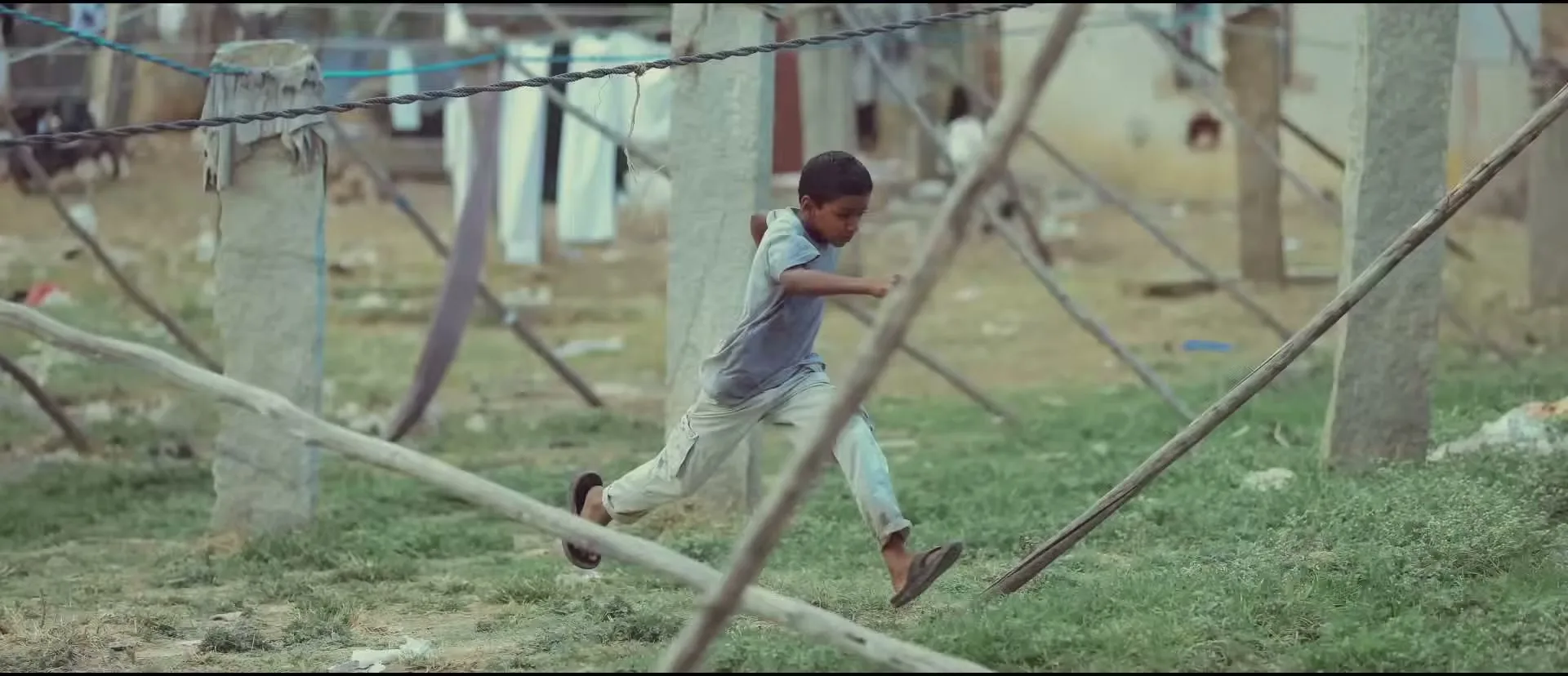 A young boy in a striped shirt and blue shorts runs barefoot on a dirt road past a stone wall and a tree with exposed roots.