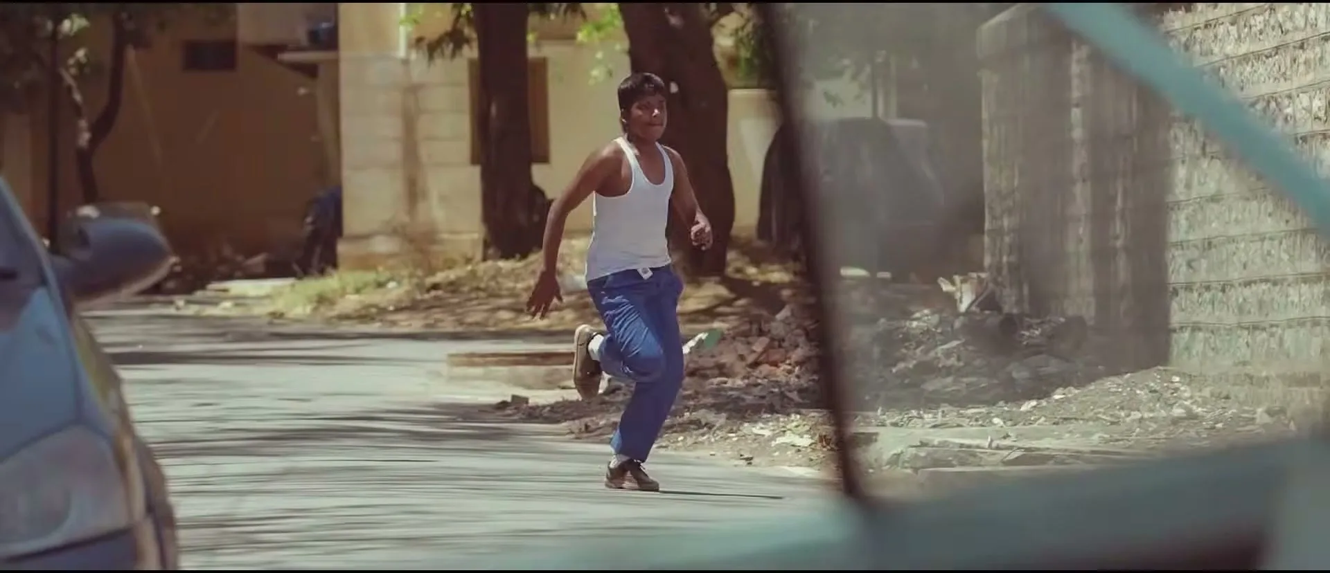 A young man in a white tank top and blue pants runs down a street, viewed through the open door of a car.