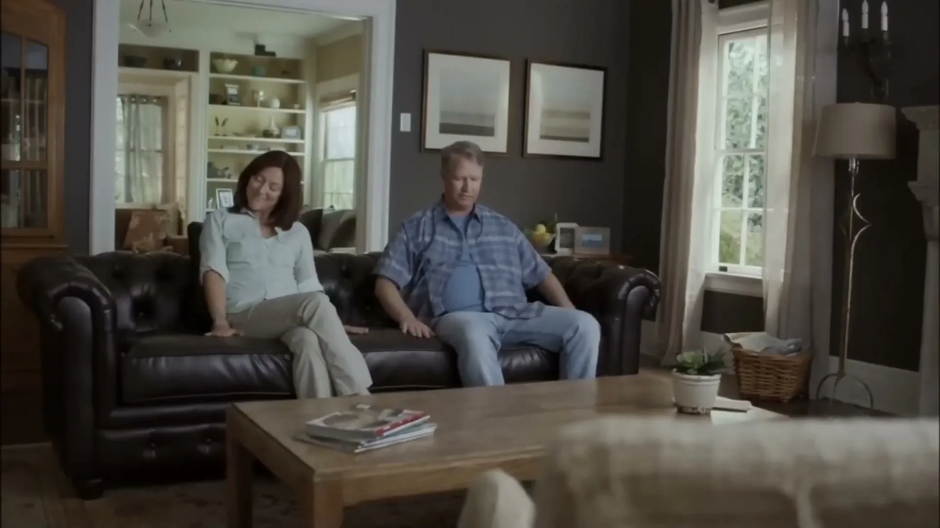 A middle-aged couple sits on a new brown leather couch in their living room, looking around contentedly.
