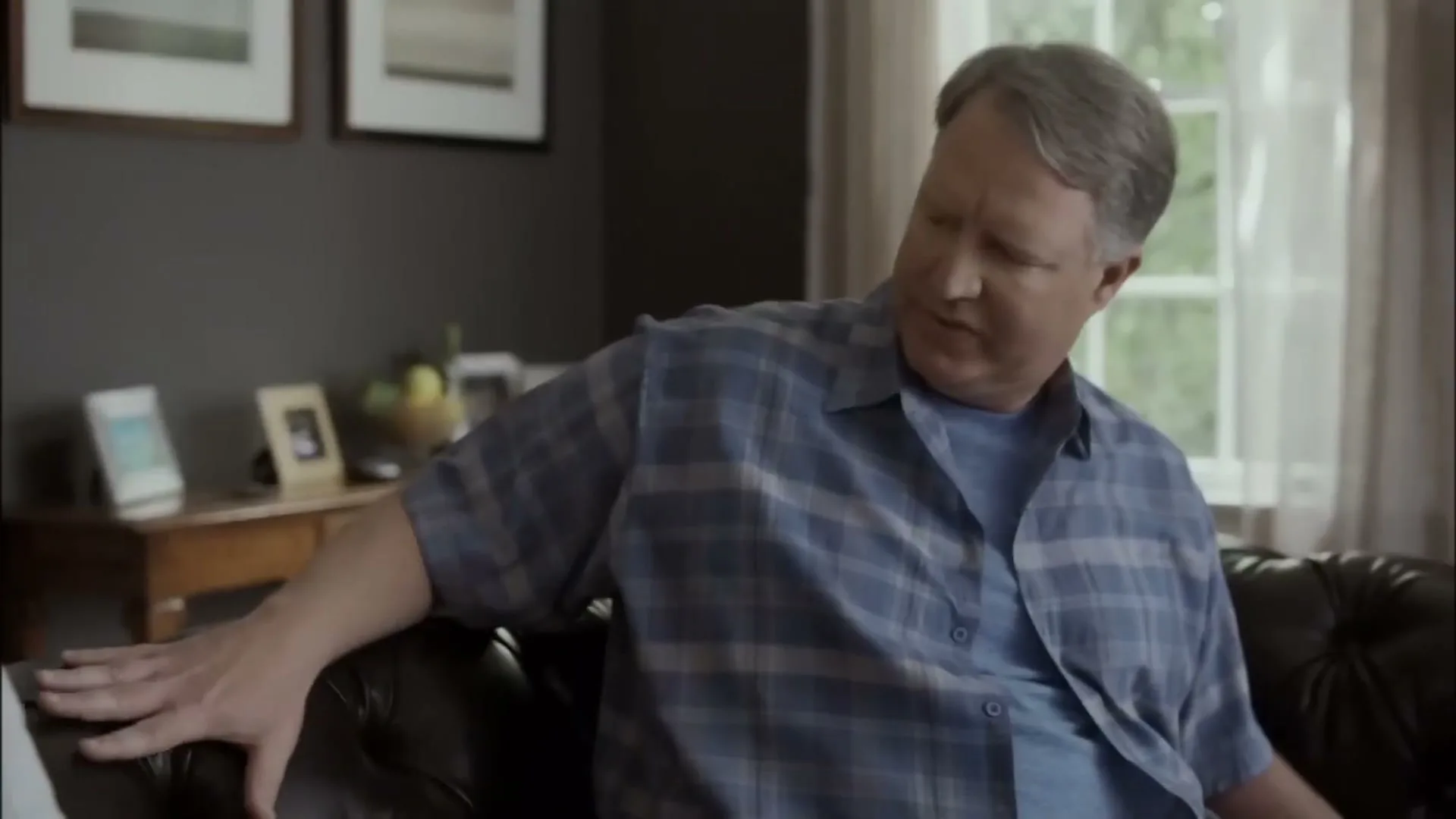 A middle-aged man in a blue plaid shirt sits on a brown leather couch, looking around a living room.