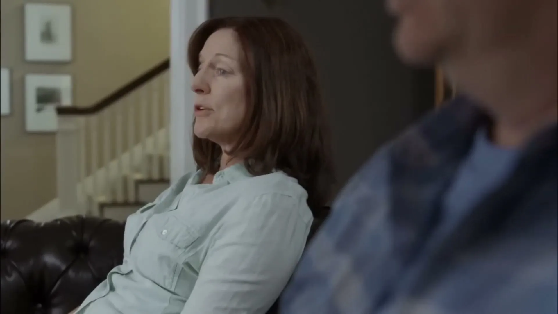 A middle-aged woman with brown hair, wearing a light blue shirt, sits on a brown leather couch, looking off-screen and talking in a living room.