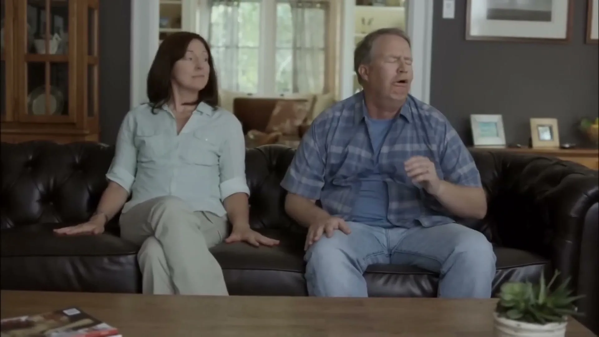 A middle-aged couple sits on a dark leather couch in their living room, looking disgusted by something off-screen.