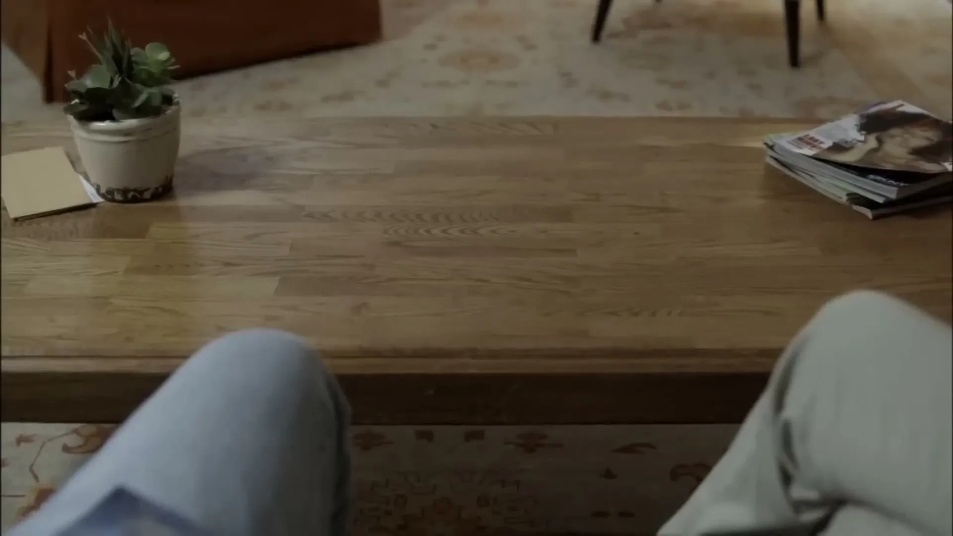 A first-person perspective shot shows a person's legs and a wooden coffee table with a plant and magazines in a living room.