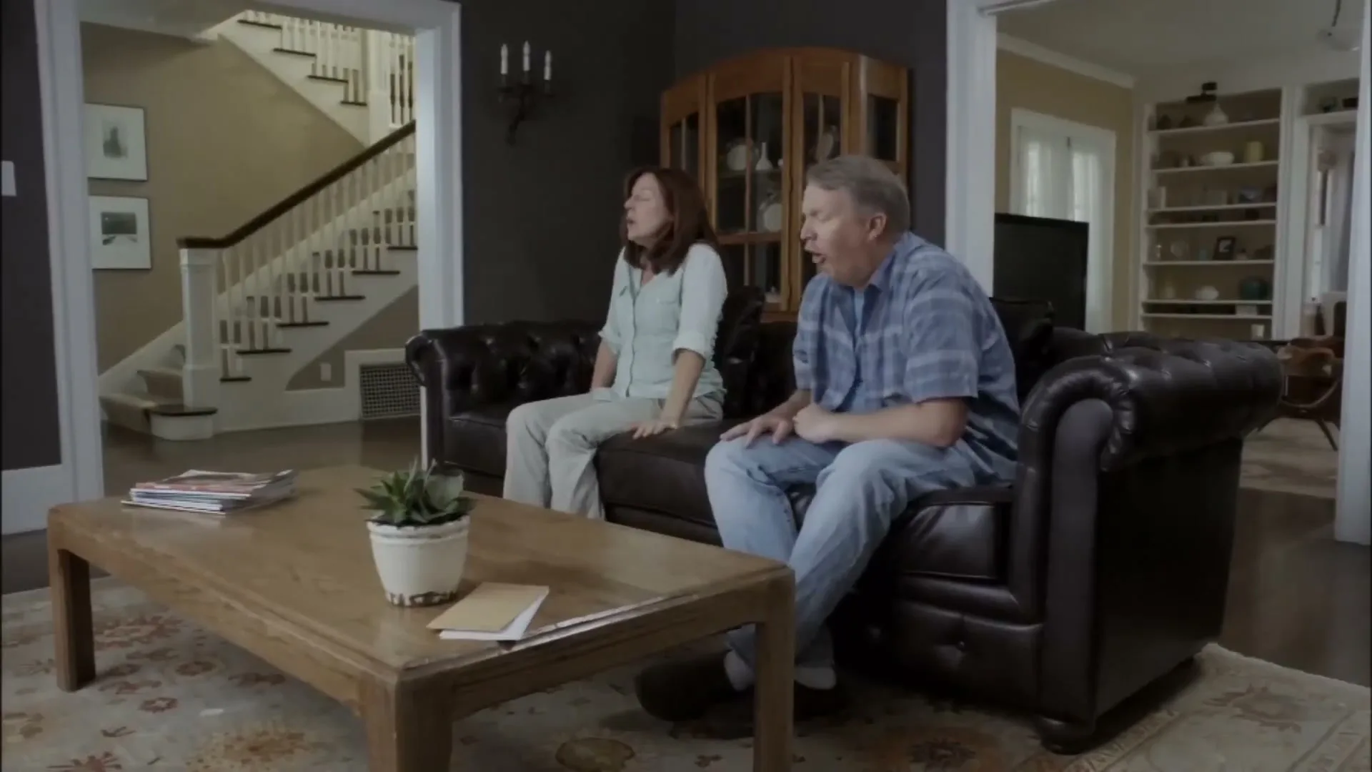 A middle-aged couple sits on a brown leather couch in their living room, looking up and reacting to something off-screen.