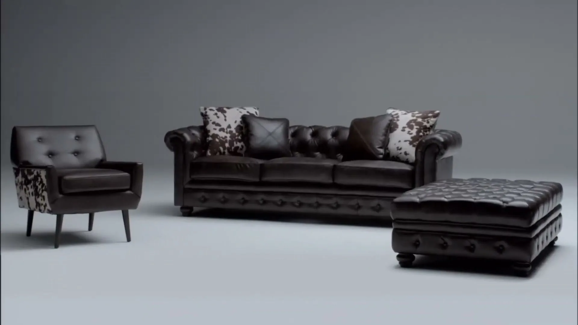 A dark brown leather sofa, armchair, and ottoman with cowhide print pillows are displayed in a studio void.