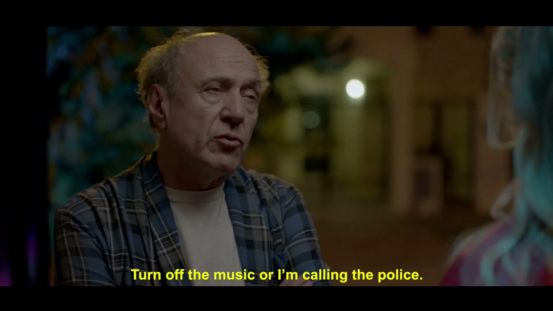 An older man with a plaid shirt and a young woman with blonde hair are talking outside at night.
