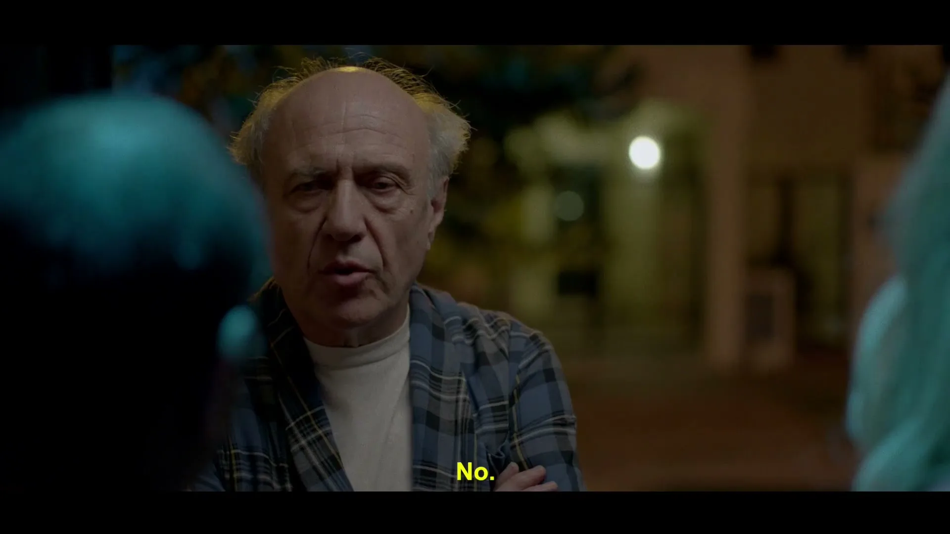 An older man with a receding hairline and a plaid shirt looks directly at the camera, speaking to someone off-screen at night.
