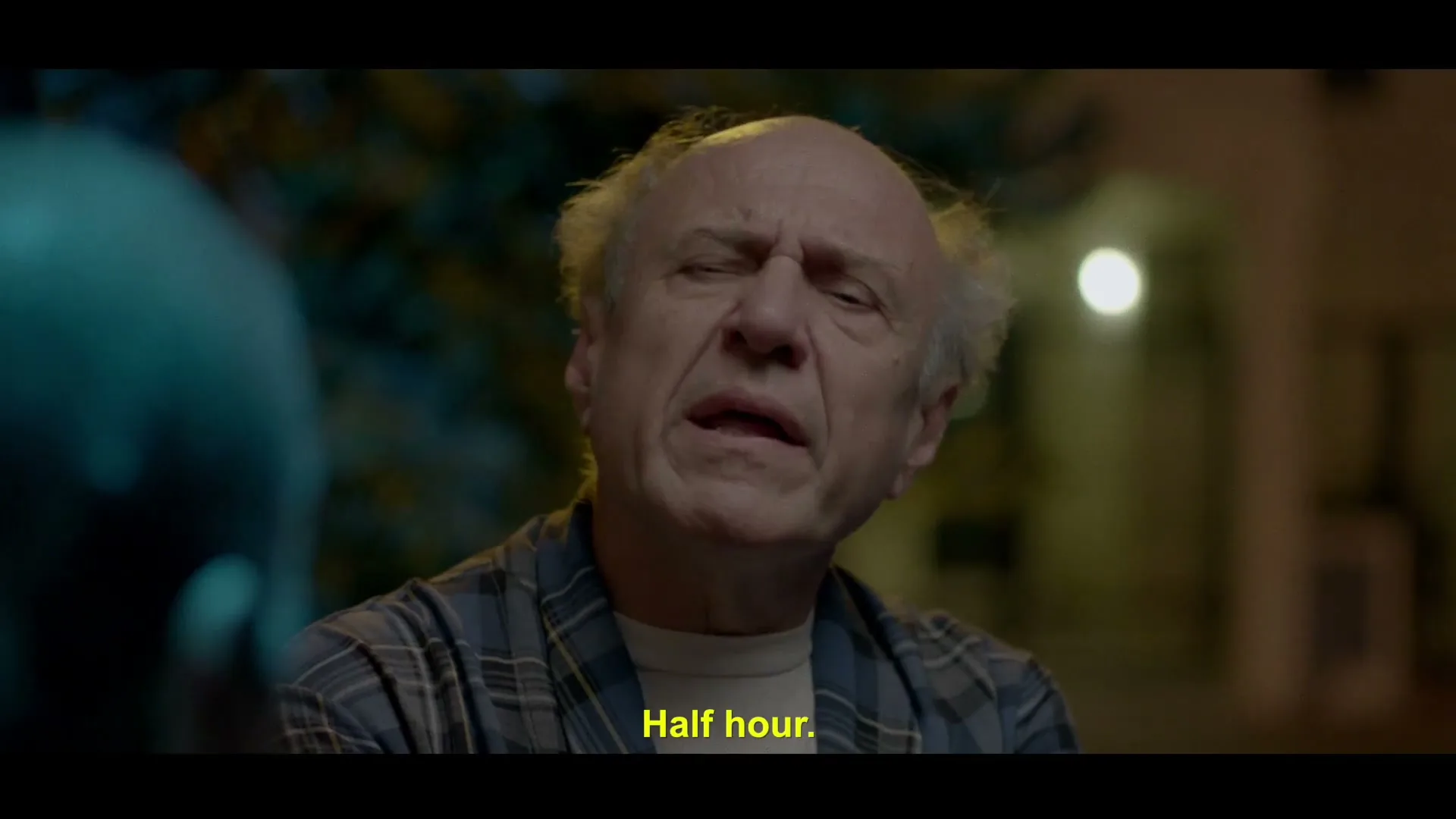 An older man with white hair and a plaid shirt looks tired and speaks to someone off-screen at night.
