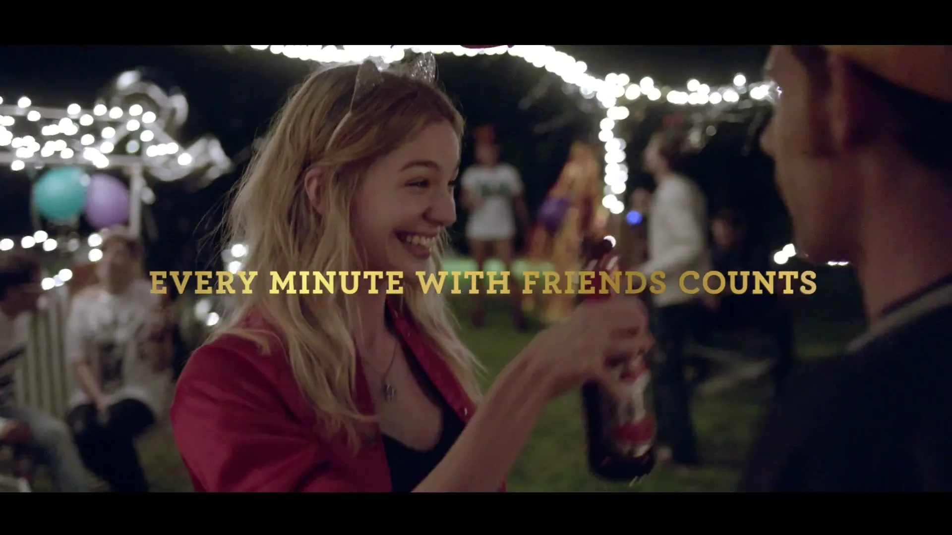 A young woman in a red jacket and cat ears smiles at a man while holding a beer bottle at a lively outdoor night party with string lights.