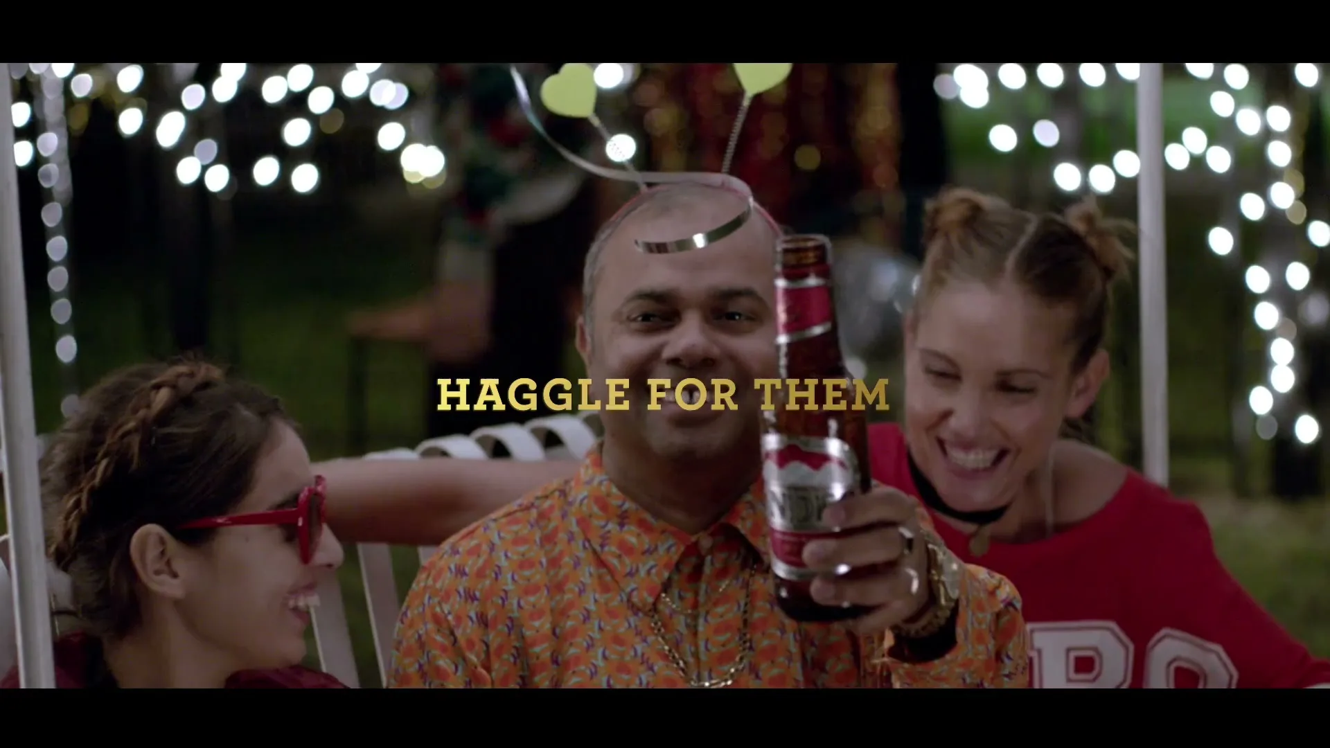A man in a patterned shirt and party headband smiles, holding up a beer bottle, flanked by two laughing women at an outdoor night party.