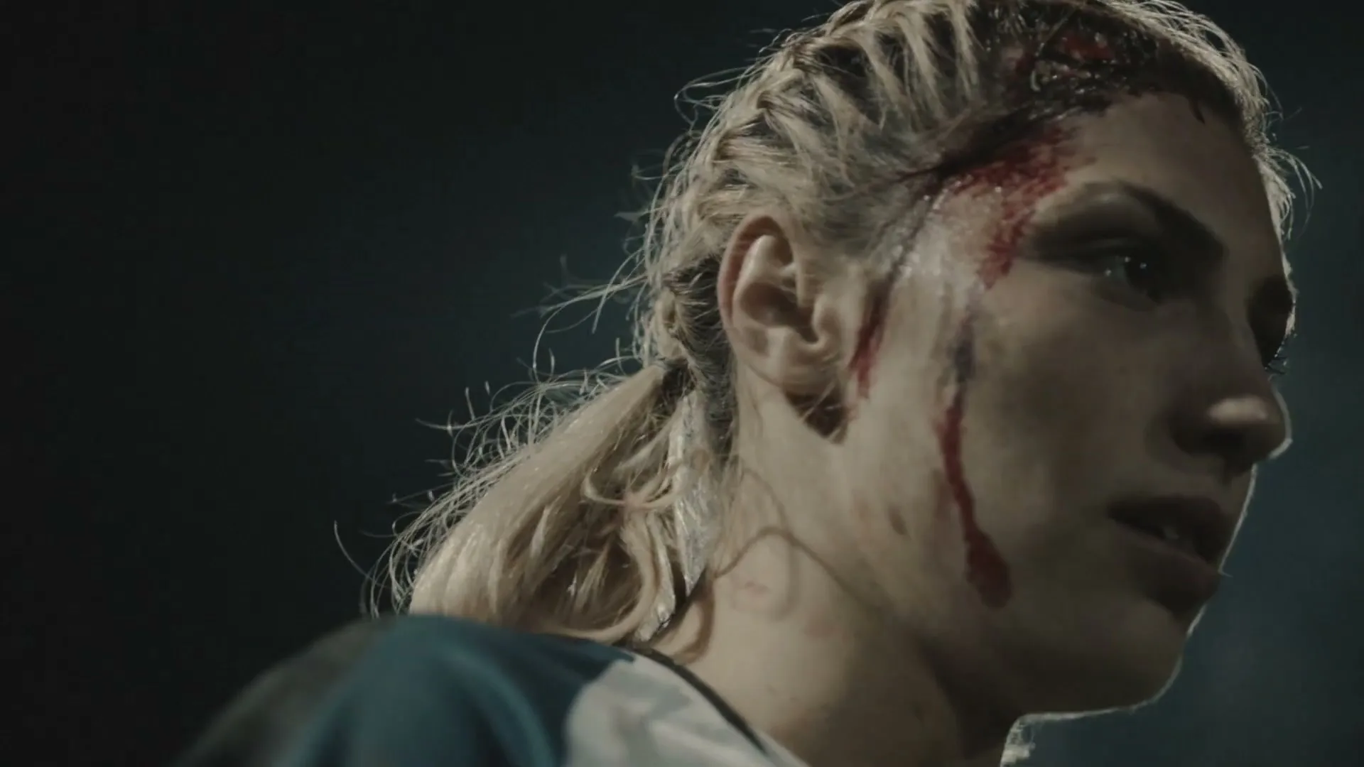 A female rugby player with braided blonde hair and blood on her face looks determined on a dark field at night.